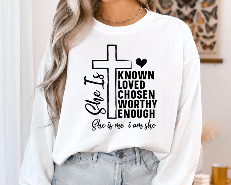 May include: White long-sleeve shirt with a black cross and the text "She Is" and "Known Loved Chosen Worthy Enough She is me i am she".