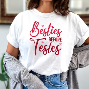 Op de afbeelding: Wit T-shirt met de tekst "Besties Before Testes" in rood met roze hartjes.