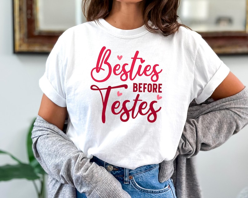 Op de afbeelding: Wit T-shirt met de tekst "Besties Before Testes" in rood met roze hartjes.