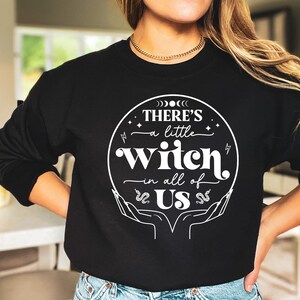 May include: Black sweatshirt with a white graphic design that says "There's a little witch in all of us" with a circle around the text and a snake design on each side of the text.