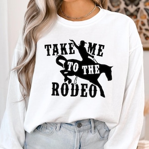 Op de afbeelding: Wit langarm shirt met zwarte tekst die "Take Me To The Rodeo" zegt en een silhouet van een persoon die een paard rijdt.
