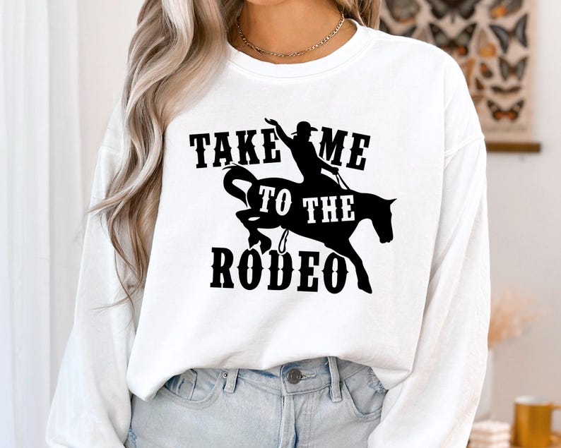 Op de afbeelding: Wit langarm shirt met zwarte tekst die "Take Me To The Rodeo" zegt en een silhouet van een persoon die een paard rijdt.