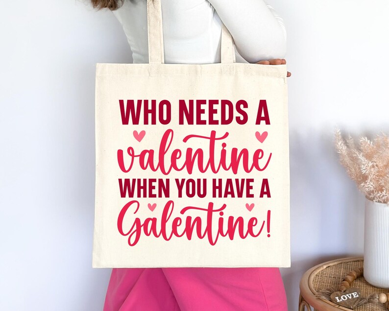 Op de afbeelding: Een witte tote bag met de tekst "Who needs a valentine when you have a Galentine!" in rode en roze letters. Er zijn kleine roze hartjes aan weerszijden van de tekst.