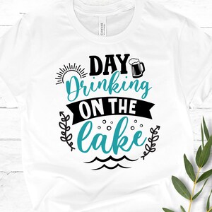 May include: White t-shirt with black text that reads "Day Drinking on the Lake" with a blue sun, a beer mug, and waves.