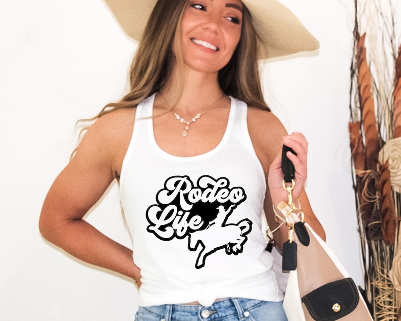 Op de afbeelding: Een wit tanktop met een zwarte afbeelding waarop "Rodeo Life" staat met een silhouet van een cowboy die een paard berijdt.