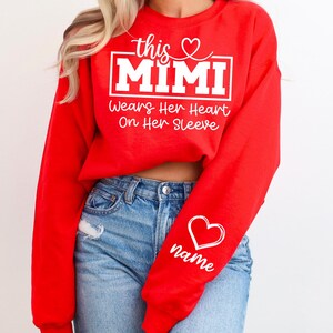 May include: A red sweatshirt with the text "this MIMI wears her heart on her sleeve" in white. A white heart with the word "name" is on the sleeve. The sweatshirt is paired with blue jeans.