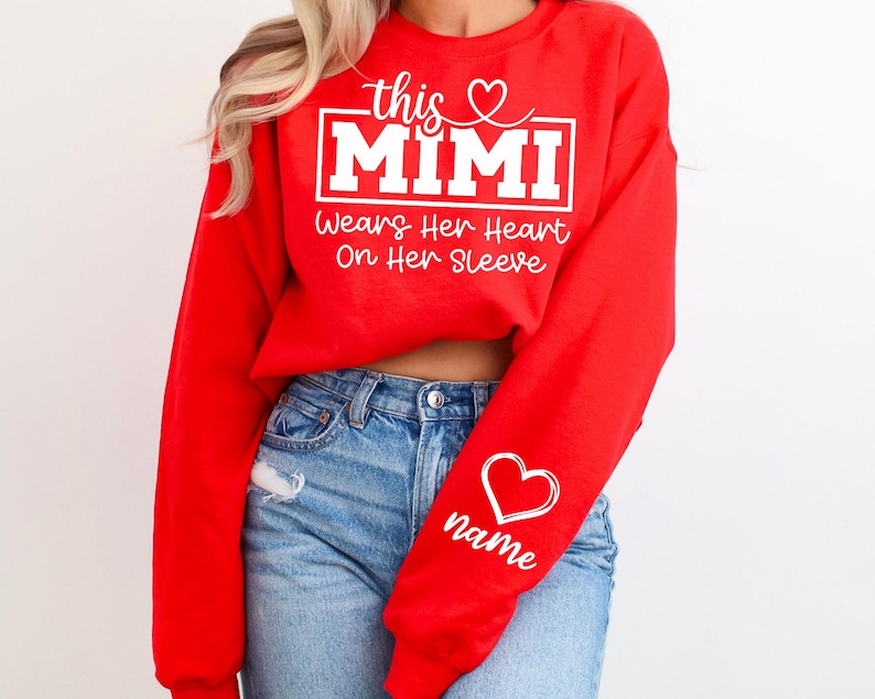 May include: A red sweatshirt with the text "this MIMI wears her heart on her sleeve" in white. A white heart with the word "name" is on the sleeve. The sweatshirt is paired with blue jeans.