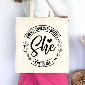 May include: A white canvas tote bag with black text that reads "Owns - Invests - Builds She - She is Me." The text is in a circle with a small heart and leaves around it.