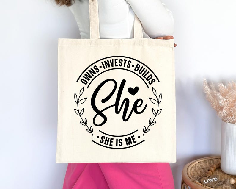 May include: A white canvas tote bag with black text that reads "Owns - Invests - Builds She - She is Me." The text is in a circle with a small heart and leaves around it.