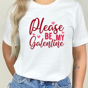 Op de afbeelding: Wit T-shirt met de tekst "Please be my Galentine" in rode letters met roze hartjes.