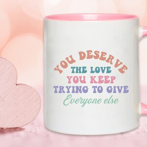 May include: White ceramic mug with a pink rim and handle. The mug has a colorful graphic that reads "You deserve the love you keep trying to give everyone else".