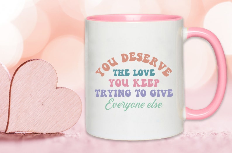 May include: White ceramic mug with a pink rim and handle. The mug has a colorful graphic that reads "You deserve the love you keep trying to give everyone else".