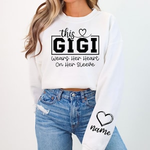 May include: White sweatshirt with the text "this GIGI Wears Her Heart On Her Sleeve" in black. A heart graphic is above the word "this" and on the sleeve, with the word "name" below it.