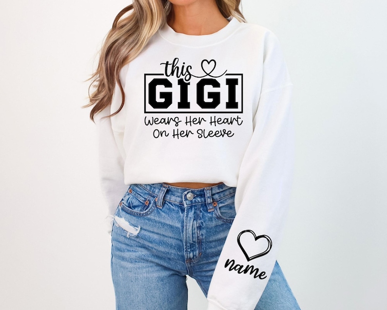 May include: White sweatshirt with the text "this GIGI Wears Her Heart On Her Sleeve" in black. A heart graphic is above the word "this" and on the sleeve, with the word "name" below it.