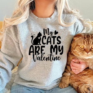 Pu&ograve; includere: Una felpa grigia con il testo "My cats are my Valentine" in lettere nere e una silhouette nera di un gatto con un cuore sul petto. Una persona indossa la felpa e tiene un gatto tabby marrone.