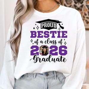 Peut inclure: Sweat-shirt blanc avec l'inscription "PROUD BESTIE of a class of 2026 Graduate" en noir et violet. Le motif comprend un chapeau de graduation et une photo d'un dipl&ocirc;m&eacute;. Le sweat-shirt est &agrave; col rond et &agrave; manches longues.