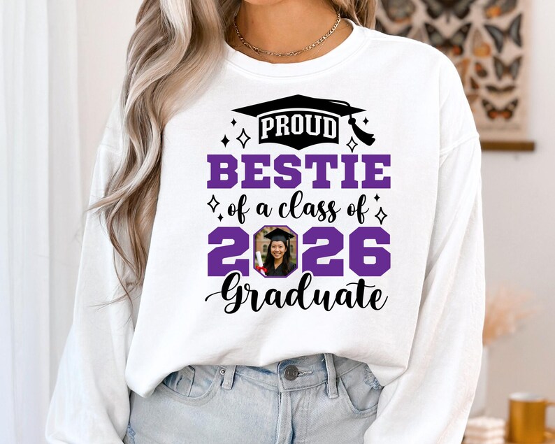 Peut inclure: Sweat-shirt blanc avec l'inscription "PROUD BESTIE of a class of 2026 Graduate" en noir et violet. Le motif comprend un chapeau de graduation et une photo d'un dipl&ocirc;m&eacute;. Le sweat-shirt est &agrave; col rond et &agrave; manches longues.
