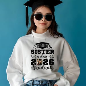 Peut inclure: Sweat-shirt blanc avec l'inscription noire "PROUD SISTER of a class of 2026 Graduate". Le sweat-shirt a un col montant et des manches longues. La personne porte une casquette de graduation et des lunettes de soleil.