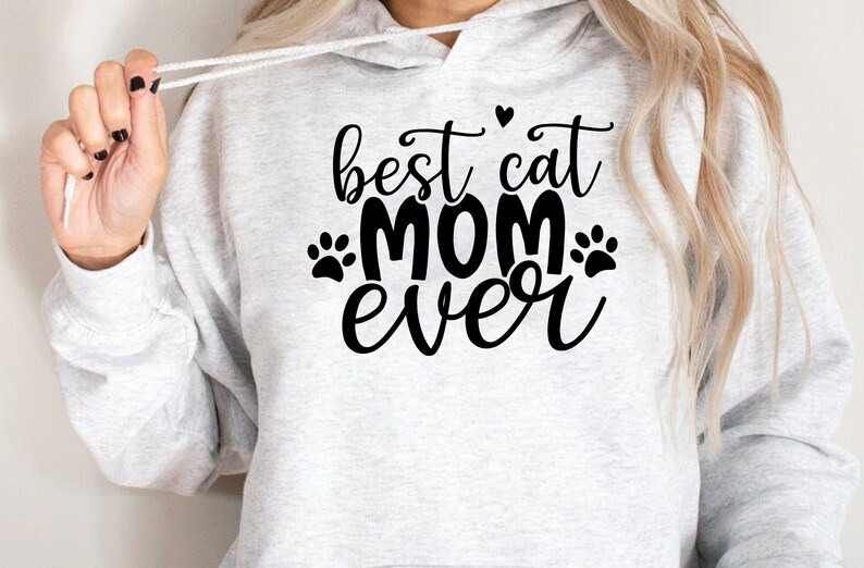 Pu&ograve; includere: Una felpa con cappuccio grigia con il testo "best cat mom ever" in lettere nere con impronte di zampe e un cuore.