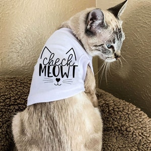 Pu&ograve; includere: Un gatto bianco che indossa una maglietta bianca con testo nero che dice "check MEOWT" e un viso di gatto nero con un naso a forma di cuore.