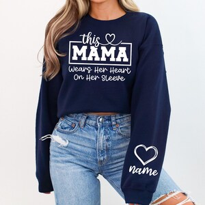 May include: Navy blue sweatshirt with white text that reads "this MAMA wears her heart on her sleeve." A white heart and the word "name" are on the sleeve. The sweatshirt is paired with blue jeans.