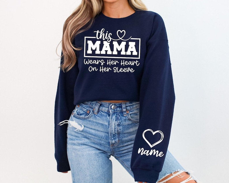 May include: Navy blue sweatshirt with white text that reads "this MAMA wears her heart on her sleeve." A white heart and the word "name" are on the sleeve. The sweatshirt is paired with blue jeans.