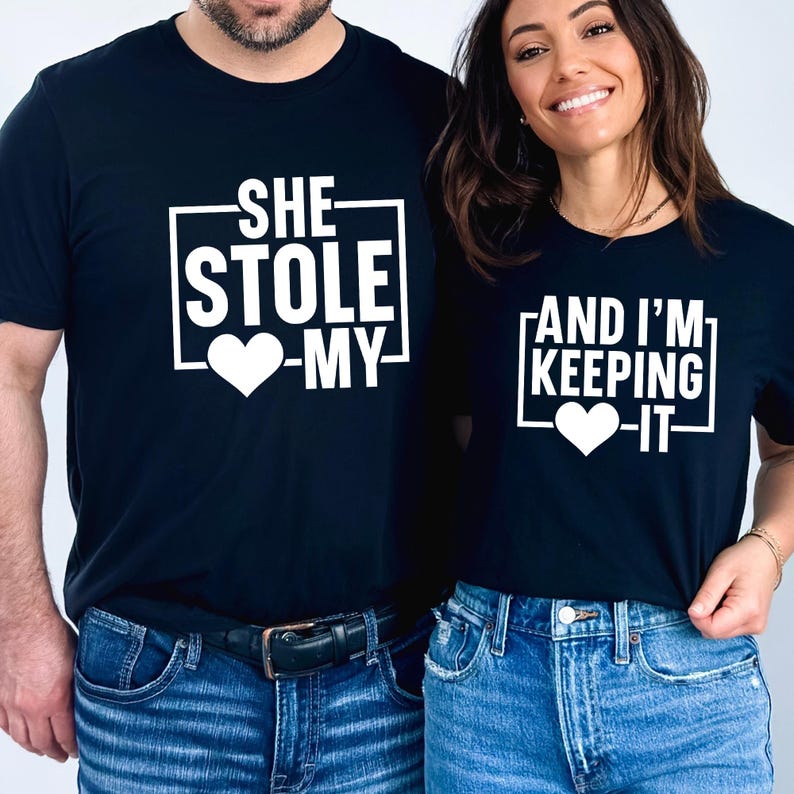 K&ouml;nnte beinhalten: Passende schwarze T-Shirts mit wei&szlig;em Text. Auf einem Shirt steht "SHE STOLE -MY-" mit einem Herz, auf dem anderen "AND I'M KEEPING -IT-" mit einem Herz. Beide Shirts werden mit blauen Jeans getragen.