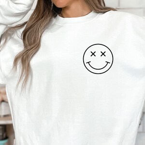 K&ouml;nnte beinhalten: Wei&szlig;er Pullover mit einem schwarzen Smiley-Motiv mit X f&uuml;r die Augen.