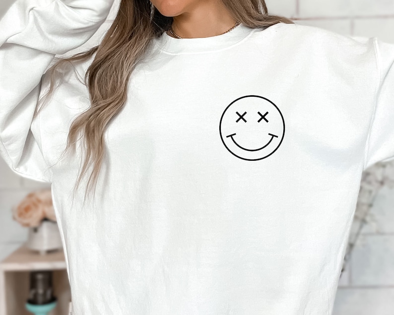 K&ouml;nnte beinhalten: Wei&szlig;er Pullover mit einem schwarzen Smiley-Motiv mit X f&uuml;r die Augen.