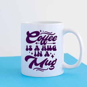 May include: White ceramic mug with a purple design that says "Coffee is a hug in a mug".