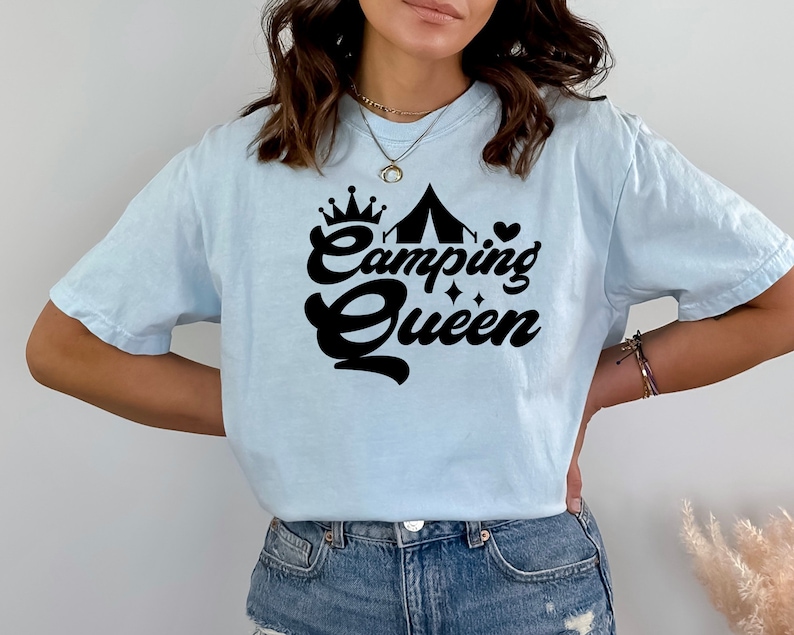 May include: A light blue t-shirt with a black graphic design that says "Camping Queen" with a crown, tent, and heart.