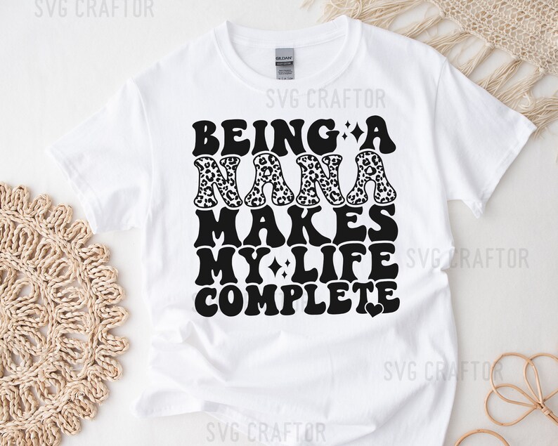 May include: White t-shirt with black text that reads "Being a Nana makes my life complete" with a heart at the end. The word "Nana" is decorated with a leopard print pattern.