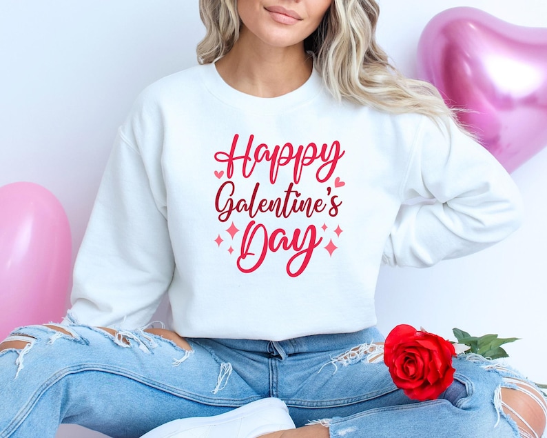 Op de afbeelding: Witte trui met de tekst "Happy Galentine's Day" in roze letters met kleine roze hartjes en glinsterende accenten.