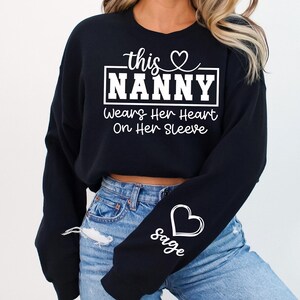 May include: Black sweatshirt with the text "this Nanny Wears Her Heart On Her Sleeve" in white. A white heart and the name "sage" are on the left sleeve. The sweatshirt is paired with blue jeans.