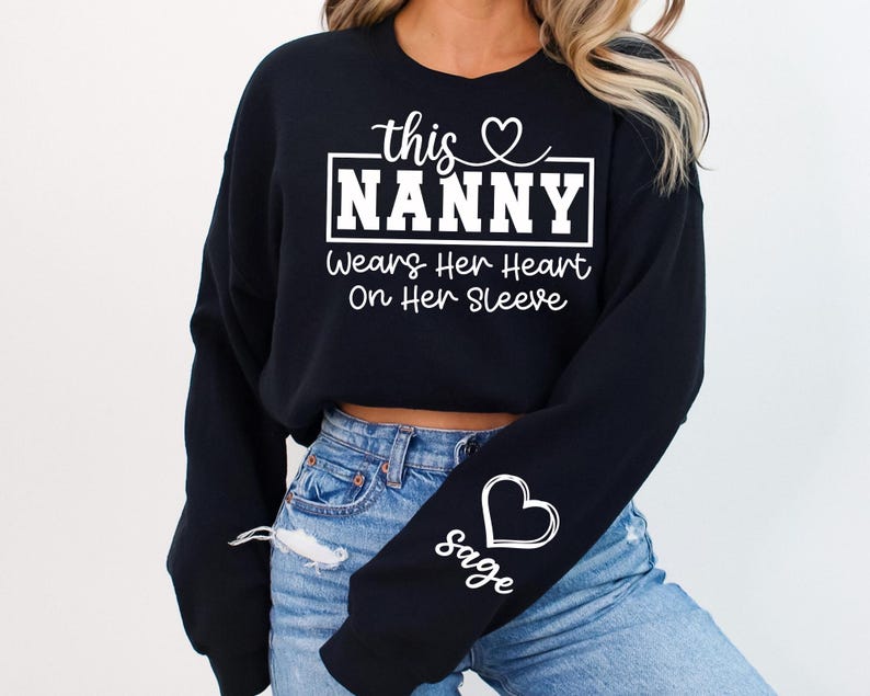 May include: Black sweatshirt with the text "this Nanny Wears Her Heart On Her Sleeve" in white. A white heart and the name "sage" are on the left sleeve. The sweatshirt is paired with blue jeans.