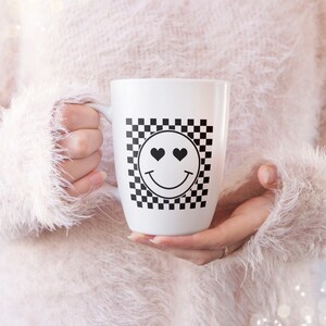 K&ouml;nnte beinhalten: Wei&szlig;e Keramiktasse mit einem schwarz-wei&szlig;en Karomuster und einem schwarzen Smiley mit Herzaugen.