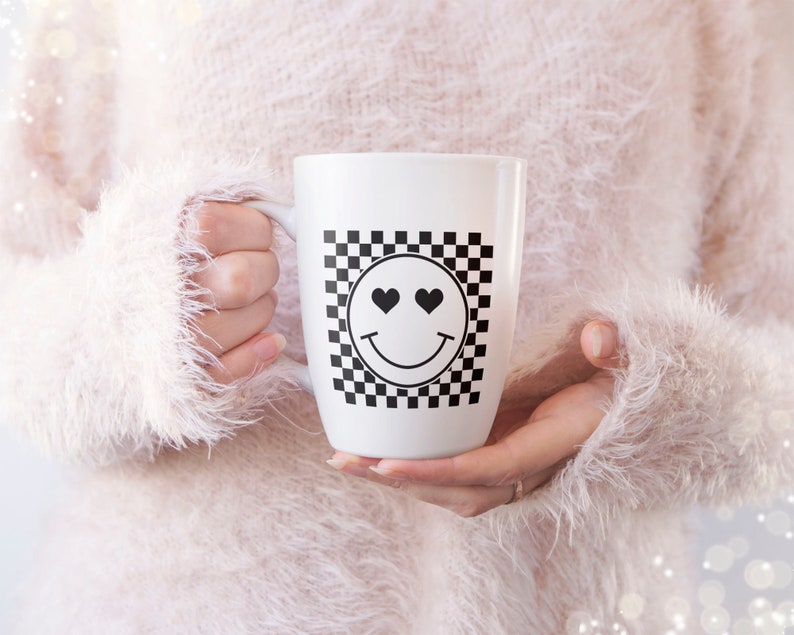 K&ouml;nnte beinhalten: Wei&szlig;e Keramiktasse mit einem schwarz-wei&szlig;en Karomuster und einem schwarzen Smiley mit Herzaugen.