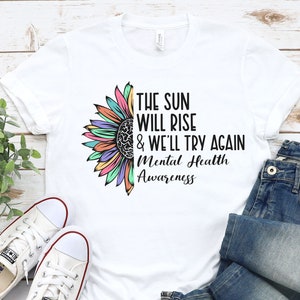 May include: White t-shirt with a colorful sunflower graphic and the text "The sun will rise & we'll try again Mental Health Awareness".