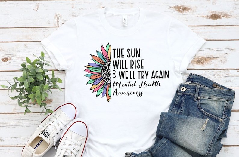 May include: White t-shirt with a colorful sunflower graphic and the text "The sun will rise & we'll try again Mental Health Awareness".