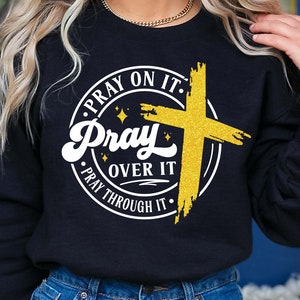 May include: Black sweatshirt with a white circle graphic featuring a gold cross and the text "Pray On It. Pray Over It. Pray Through It."