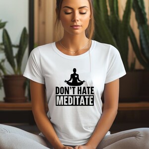 May include: A white t-shirt with a black silhouette of a person meditating with a heart in their chest. The text "Don't Hate Meditate" is printed below the silhouette.