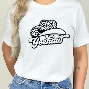 Op de afbeelding: Wit T-shirt met een zwart-witte afbeelding van een cowboyhoed met luipaardprint en het woord "Yeehaw" in een retro-lettertype.