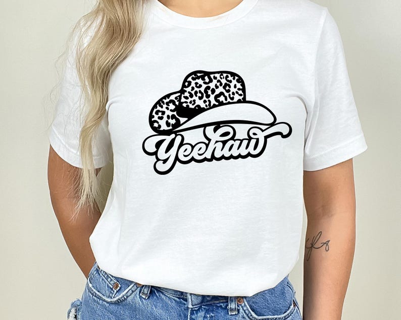 Op de afbeelding: Wit T-shirt met een zwart-witte afbeelding van een cowboyhoed met luipaardprint en het woord "Yeehaw" in een retro-lettertype.