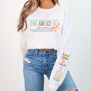 May include: White sweatshirt with the text "HIS GRACE IS enough" in rainbow colors and "2 CORINTHIANS 12:9" in black text. The right sleeve has the text "GOD is LOVE" in rainbow colors with a pink heart.