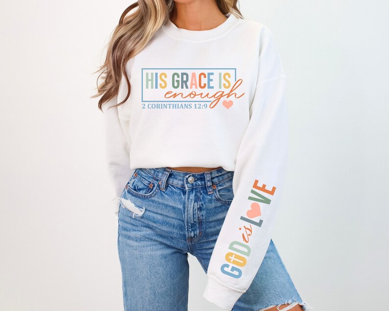 May include: White sweatshirt with the text "HIS GRACE IS enough" in rainbow colors and "2 CORINTHIANS 12:9" in black text. The right sleeve has the text "GOD is LOVE" in rainbow colors with a pink heart.