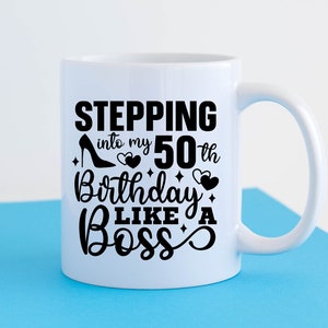 May include: White ceramic mug with a black design that says "Stepping into my 50th Birthday Like a Boss" with a high heel shoe and hearts.