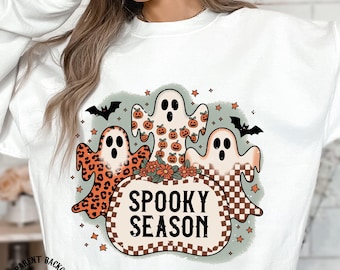 May include: White sweatshirt featuring a Halloween-themed graphic. The design includes three cartoon ghosts, pumpkins, bats, and the text "Spooky Season" in a pumpkin-shaped frame. The image is a PNG file, 14/14 inches.