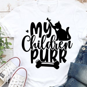 Pu&ograve; includere: Maglietta bianca con testo nero che recita "I miei figli fanno le fusa" e una silhouette nera di un gatto che gioca con un topo giocattolo. Un grafico di un osso &egrave; sotto il testo.