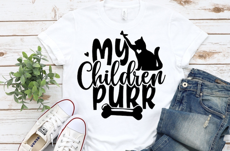 Pu&ograve; includere: Maglietta bianca con testo nero che recita "I miei figli fanno le fusa" e una silhouette nera di un gatto che gioca con un topo giocattolo. Un grafico di un osso &egrave; sotto il testo.