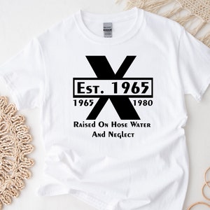 May include: White t-shirt with a black graphic design. The graphic features a large black X with the text "Est. 1965" and "1965" and "1980" below the X. The text "Raised on Hose Water and Neglect" is below the graphic.